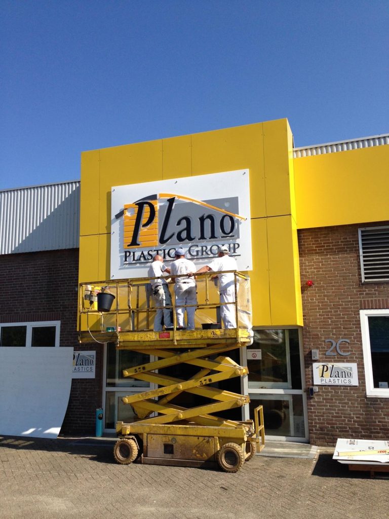 Medewerkers op een hoogwerker monteren het Plano Plastics Group-bord op de gevel aan de Hurksestraat